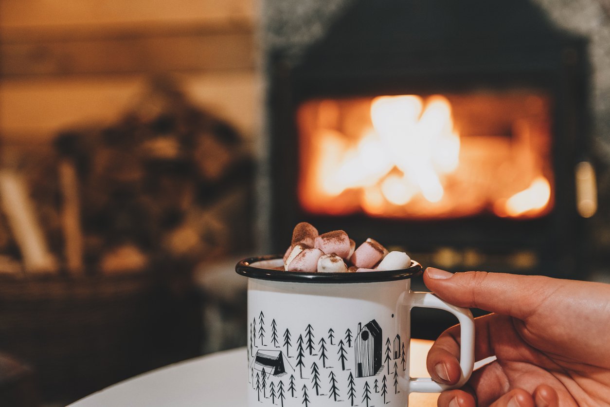 Woman's-Hand-Holding-Cup-Of-Hot-Chocolate-with-Marshmallows-In-Front-Of-Fireplace-In-Cosy-Cabin