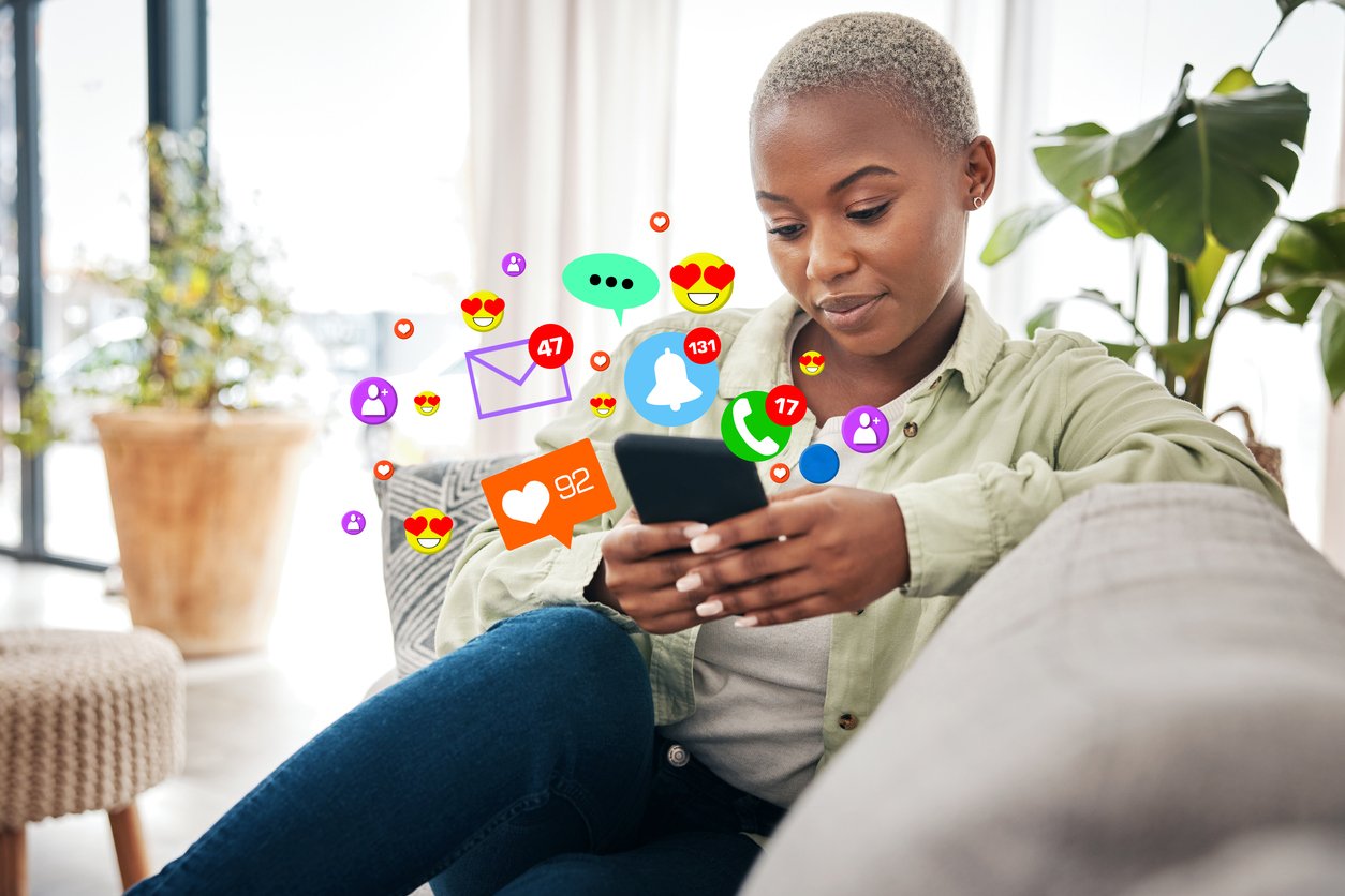 Woman-on-sofa-at-home-with-phone-social-media-icons-and-emojis-surround-the-phone.