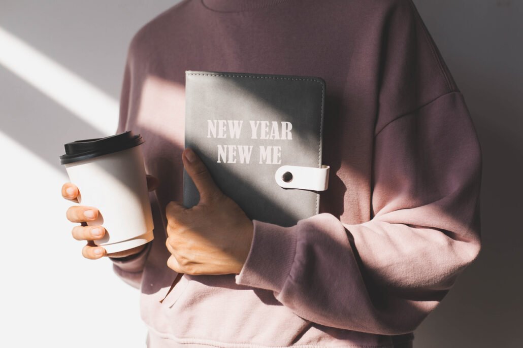 female-hand-holding-a-diary-with-the-text-new-year-new-me-and-paper-cup-with-hot-drink-on-the-background-of-a-pink-sweater-concept-of-planning-goals-and-plans-for-the-new-year-motivation.