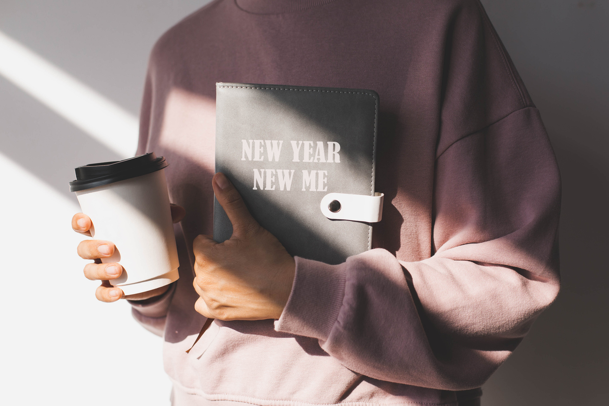 female-hand-holding-a-diary-with-the-text-new-year-new-me-and-paper-cup-with-hot-drink-on-the-background-of-a-pink-sweater-concept-of-planning-goals-and-plans-for-the-new-year-motivation.
