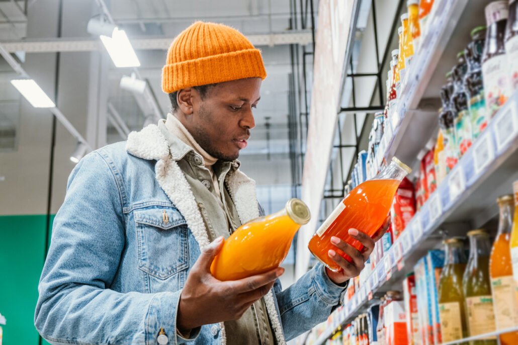 Millennial-man-in-a-stylish-orange-hat-and-denim-jacket-chooses-natural-juice-in-glass-bottles-in-a-grocery-supermarket-standing-at-the-shelves-with-products