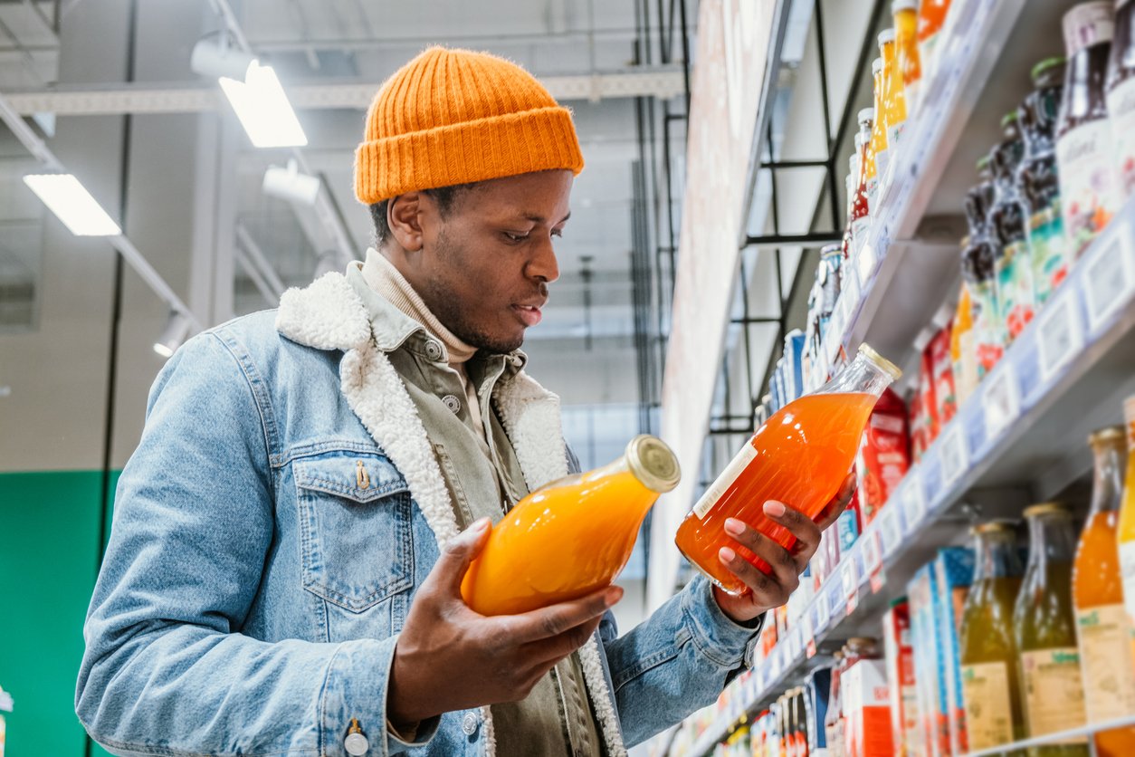 Millennial-man-in-a-stylish-orange-hat-and-denim-jacket-chooses-natural-juice-in-glass-bottles-in-a-grocery-supermarket-standing-at-the-shelves-with-products