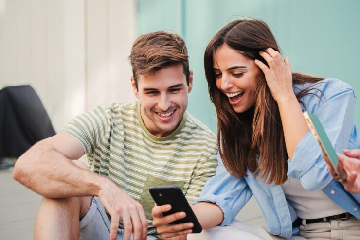 Two-happy-friends-watching-funny-videos-browsing-on-internet-app with a smartphone and smiling together. Caucasian couple using a cell phone to post in a social media and chatting online.
